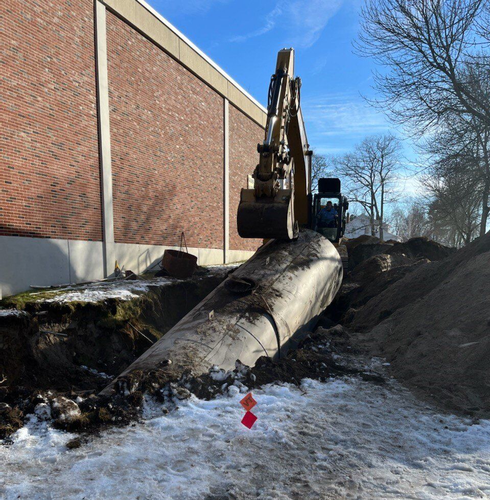 Excavator lifting an underground storage tank out of a snowy excavation beside a brick building