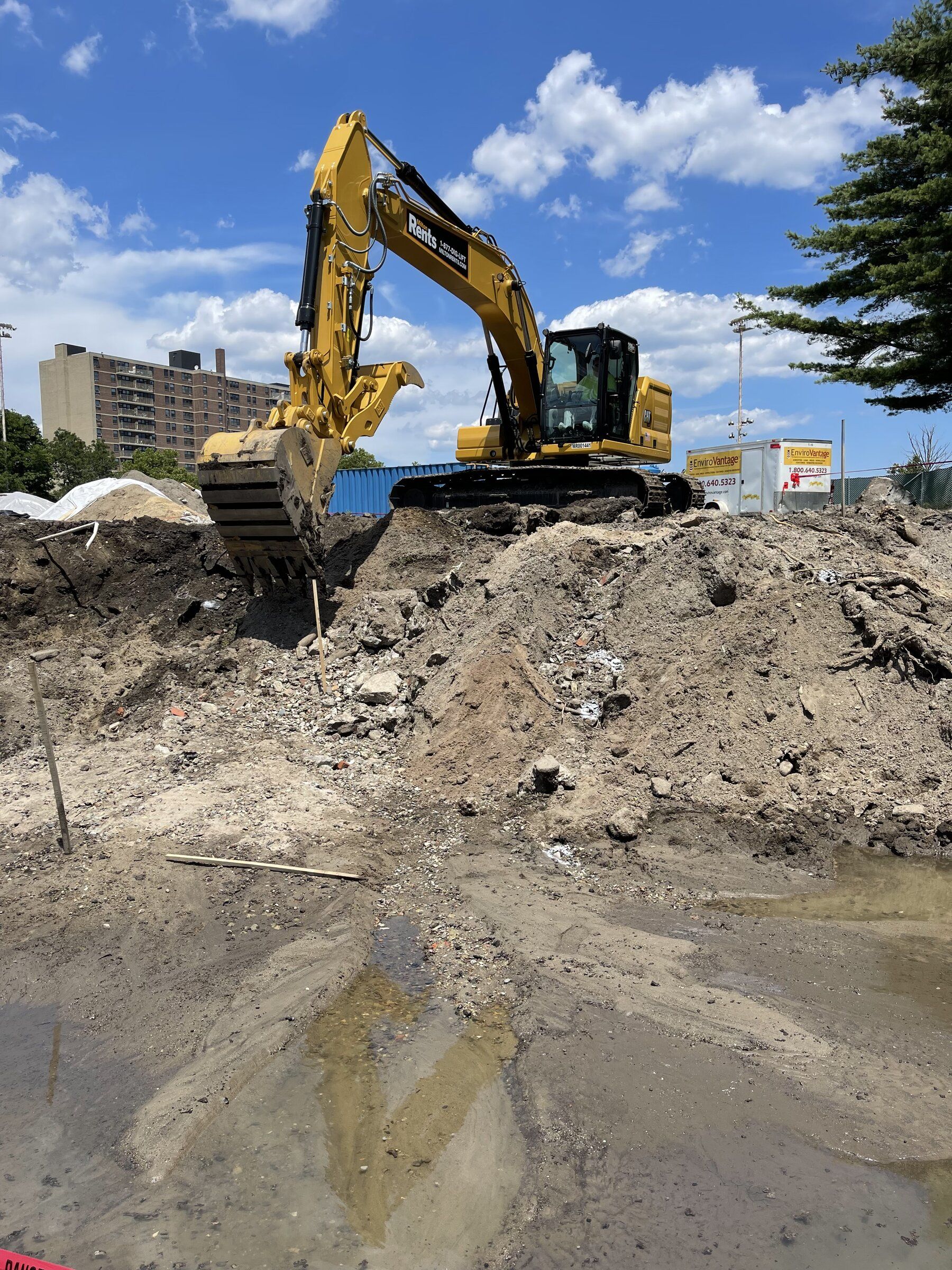 Excavator on a stockpile of impacted soil next to a water-filled excavation