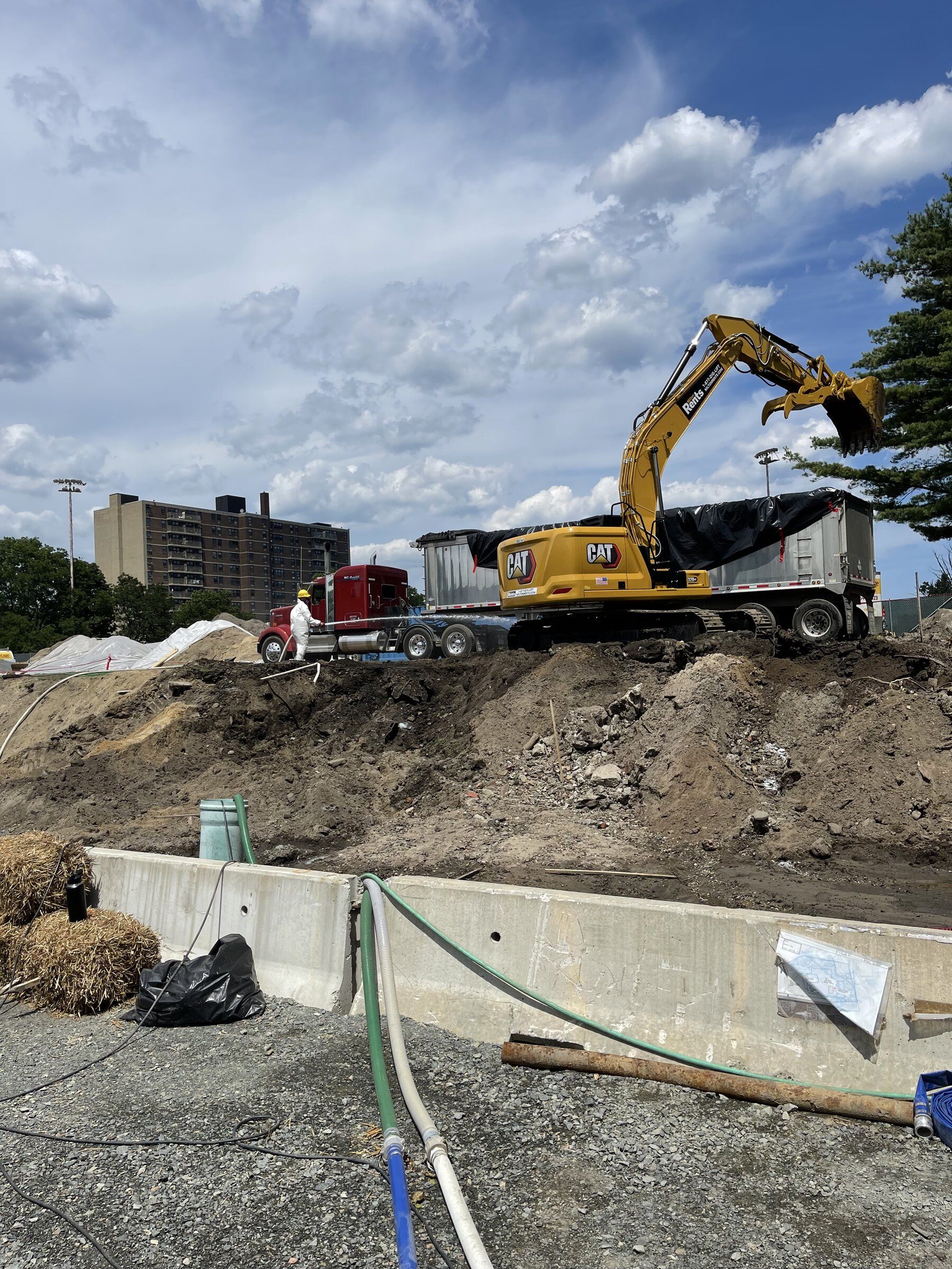 Excavator loading a dump trailer behind a concrete barrier on a cleared lot