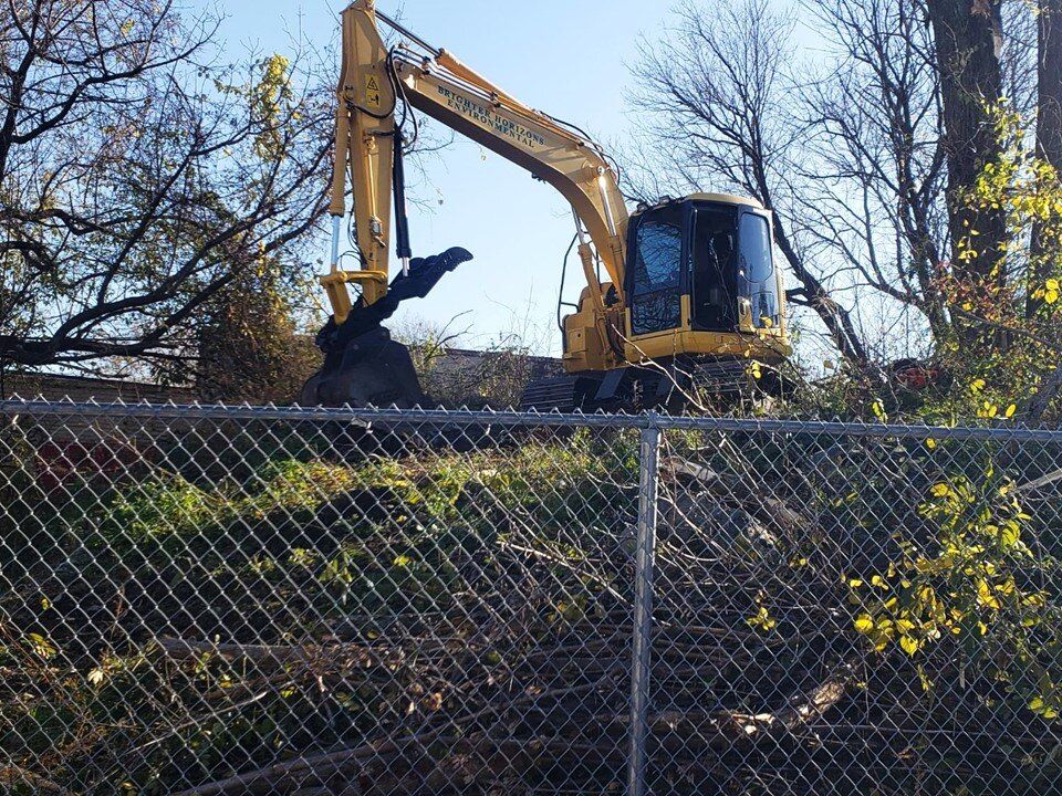 Excavator clearing an overgrown lot behind a chain-link fence
